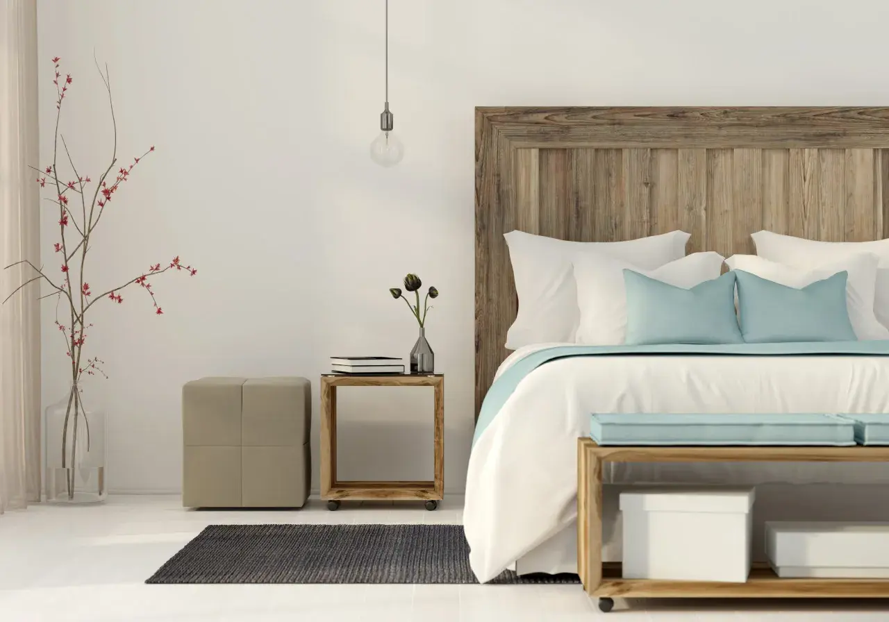 Un letto feng shui con testiera in legno grezzo, cuscini azzurri e bianchi, un vaso con rami rossi e un pouf beige.