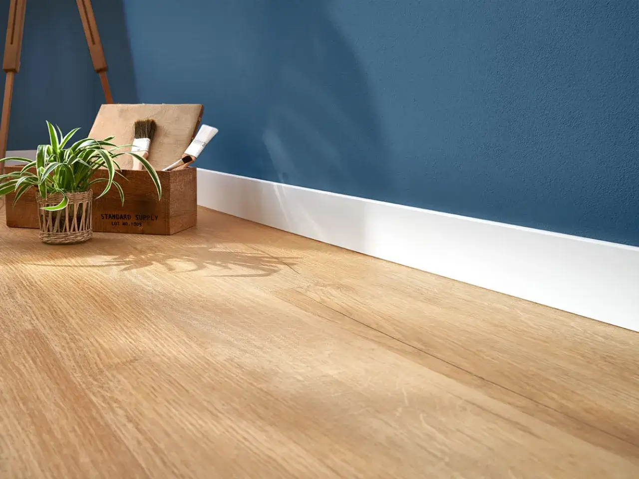Un angolo di casa con un pratico pavimento laminato effetto legno, una pianta e una cassetta con pennelli.