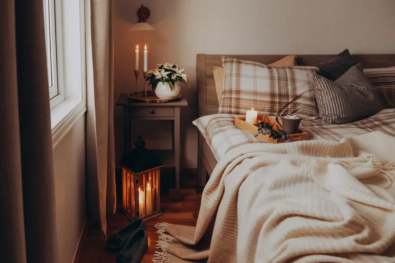 Accogliente camera da letto colori caldi con candele accese, coperta morbida e un vassoio con una tazza fumante.