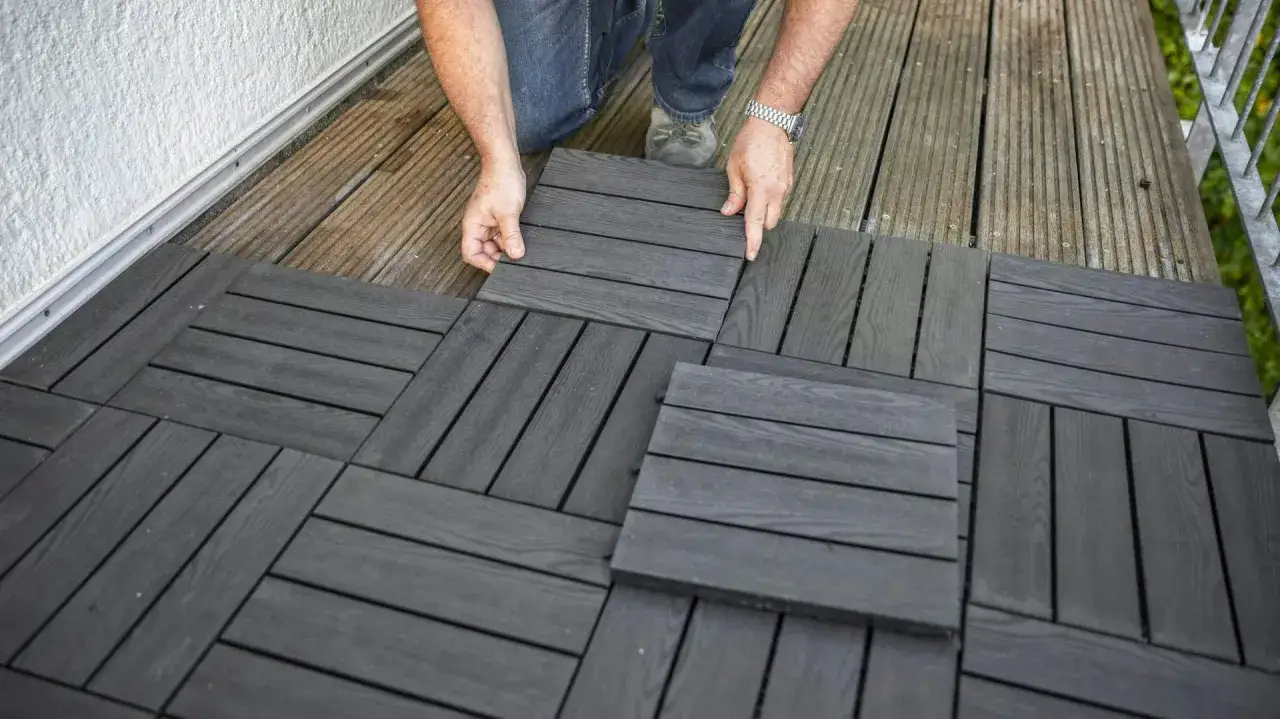 Mani che posano le nuove piastrelle per pavimento di balcone grigio scuro su una vecchia pavimentazione in legno.