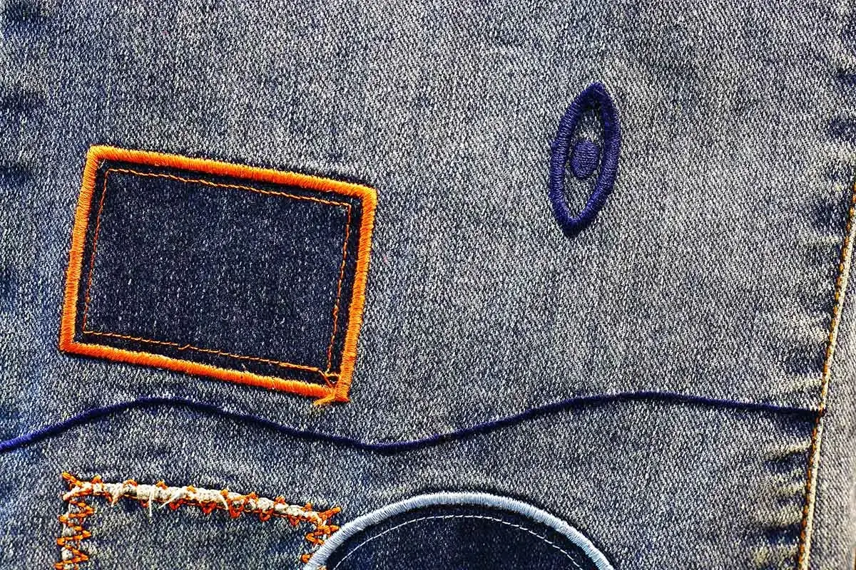Dettaglio di jeans con toppe quadrate e circolari, un occhio ricamato e cuciture decorative. Un esempio di come mettere le toppe ai pantaloni per personalizzarli.