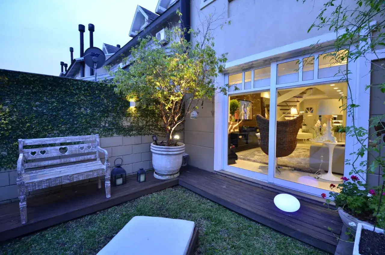 Giardino con panca in legno e albero illuminato, vista su un soggiorno accogliente attraverso vetrata. Il piano terra poco luminoso si apre su uno spazio esterno curato.