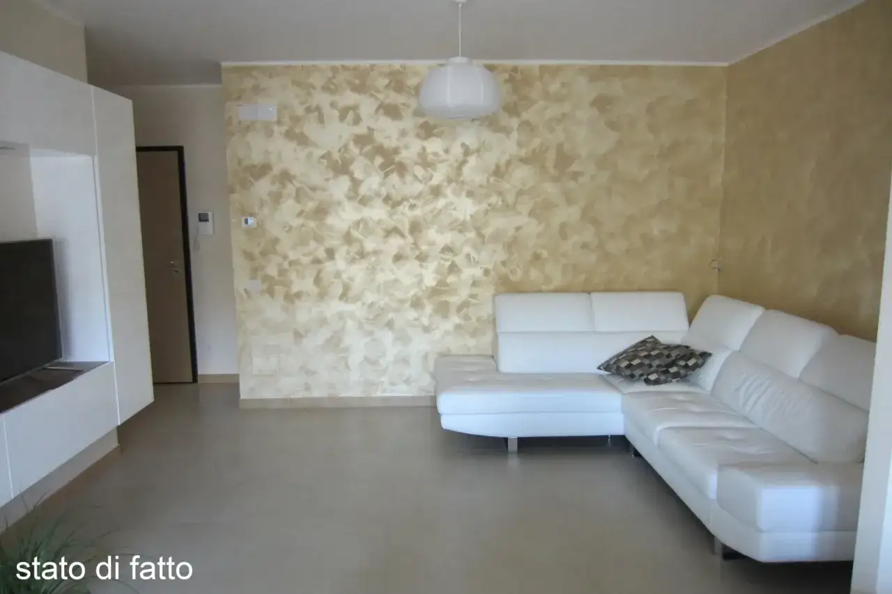 Soggiorno con pareti soggiorno due colori: una con effetto materico beige e oro, l'altra color sabbia. Divano bianco angolare.