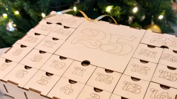 Calendario dell'avvento fatto a mano in legno, con numeri incisi e piccoli cassetti, sullo sfondo di un albero di Natale illuminato.