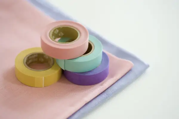 Rotoli di nastro adesivo per tessuti colorati (giallo, rosa, verde acqua, viola) su stoffe pastello. Utile per decorare o per un uso pratico su tessuti.