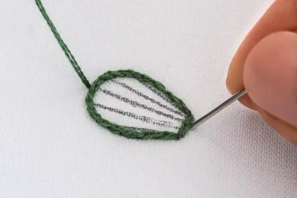 Dettaglio di schemi ricamo punto raso su tessuto bianco. Un ago con filo verde sta completando il contorno di una foglia.