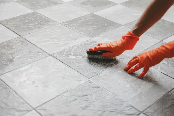 Mani con guanti arancioni che puliscono le fughe delle piastrelle con una spazzola, creando schiuma.