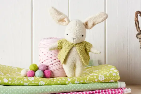 Coniglietto di stoffa con giacca verde, accanto a gomitolo rosa e palline colorate. Un cartamodello coniglio stoffa per creare il tuo amico.