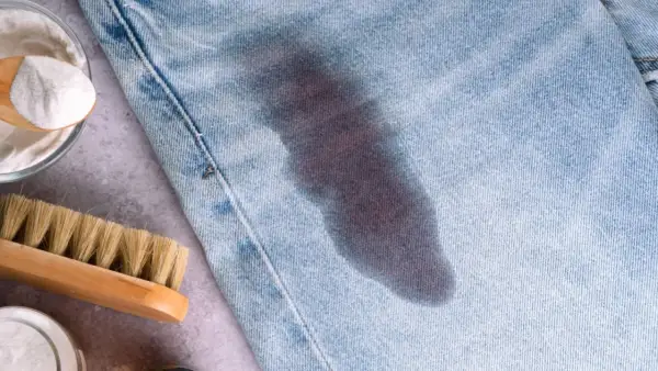 Un jeans con macchie di grasso scure. Accanto, una spazzola e una ciotola con bicarbonato, ingredienti per rimuovere le macchie.