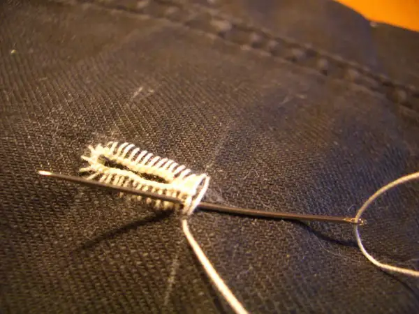 Un ago con filo bianco crea un punto occhiello a mano su tessuto scuro, un dettaglio di sartoria meticoloso.