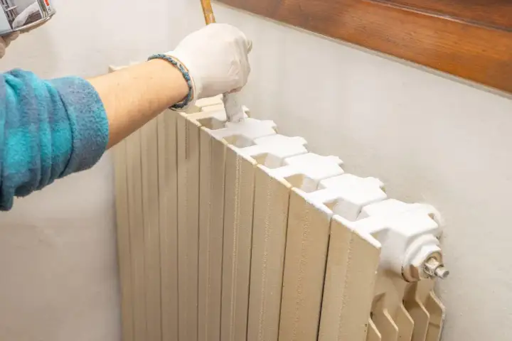 Mano con guanto bianco che applica vernici per termosifoni con un pennello. Il radiatore è beige e la vernice è bianca.
